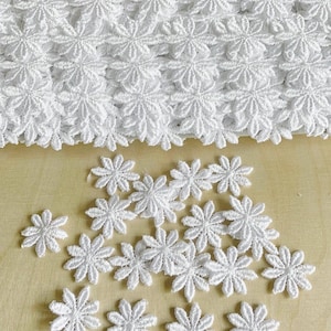 Puede incluir: Ribete de encaje blanco con flores de margarita y aplicaciones individuales de margarita. El ribete está dispuesto en filas, y las aplicaciones están dispersas sobre una superficie marrón claro. La tela es delicada.