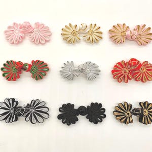 Puede incluir: Nueve juegos de cierres de rana decorativos en varios colores, incluyendo rosa, dorado, rojo, plateado, negro y combinaciones. Cada cierre presenta un diseño floral, adecuado para adornar prendas o manualidades.