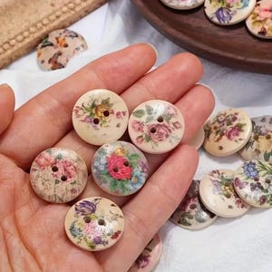 May include: A selection of round wooden buttons, each decorated with a floral design. The buttons display a variety of colourful flower patterns, including roses, on a cream backdrop. Each button has two holes for sewing.