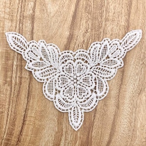 May include: A white lace applique with a floral design. The symmetrical applique features a central flower motif and intricate patterns. The delicate lace is set against a wooden background.