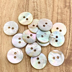 May include: A selection of iridescent, round buttons, each with two holes. The buttons have a pearlescent sheen, showing colours like white, pink, and blue. They are arranged on a wooden surface.