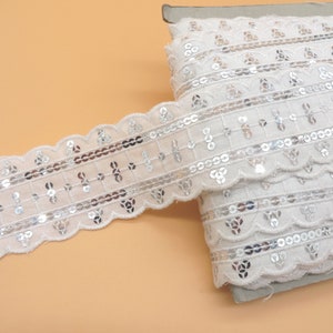 Op de afbeelding: Witte kantband met zilveren pailletten. Het band heeft een gegolfde rand en is versierd met een herhalend patroon van pailletten en borduurwerk.