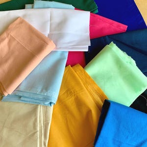 May include: A collection of folded fabric pieces in various solid colours including white, peach, light blue, cream, yellow, lime green, dark green, royal blue, navy blue, magenta, and dark blue, arranged in an overlapping, layered composition.