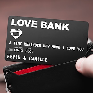 May include: A black metal card with the words "LOVE BANK" printed in white. The card has a small heart icon and the text "A TINY REMINDER HOW MUCH I LOVE YOU". It also includes the text "VALID FROM 08/13 2004 KEVIN & CAMILLE".