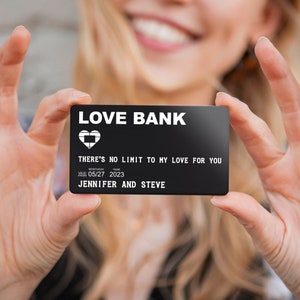 May include: A black card with white text that reads "LOVE BANK" and "THERE'S NO LIMIT TO MY LOVE FOR YOU". The card also includes the date "05/27 2023" and the names "JENNIFER AND STEVE".