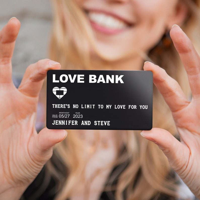 May include: A black card with white text that reads "LOVE BANK" and "THERE'S NO LIMIT TO MY LOVE FOR YOU". The card also includes the date "05/27 2023" and the names "JENNIFER AND STEVE".