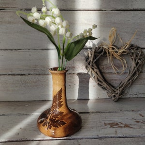 May include: A brown ceramic vase with a fern design contains white lily of the valley flowers. A woven heart decoration is to the right. The vase is on a white wooden surface.