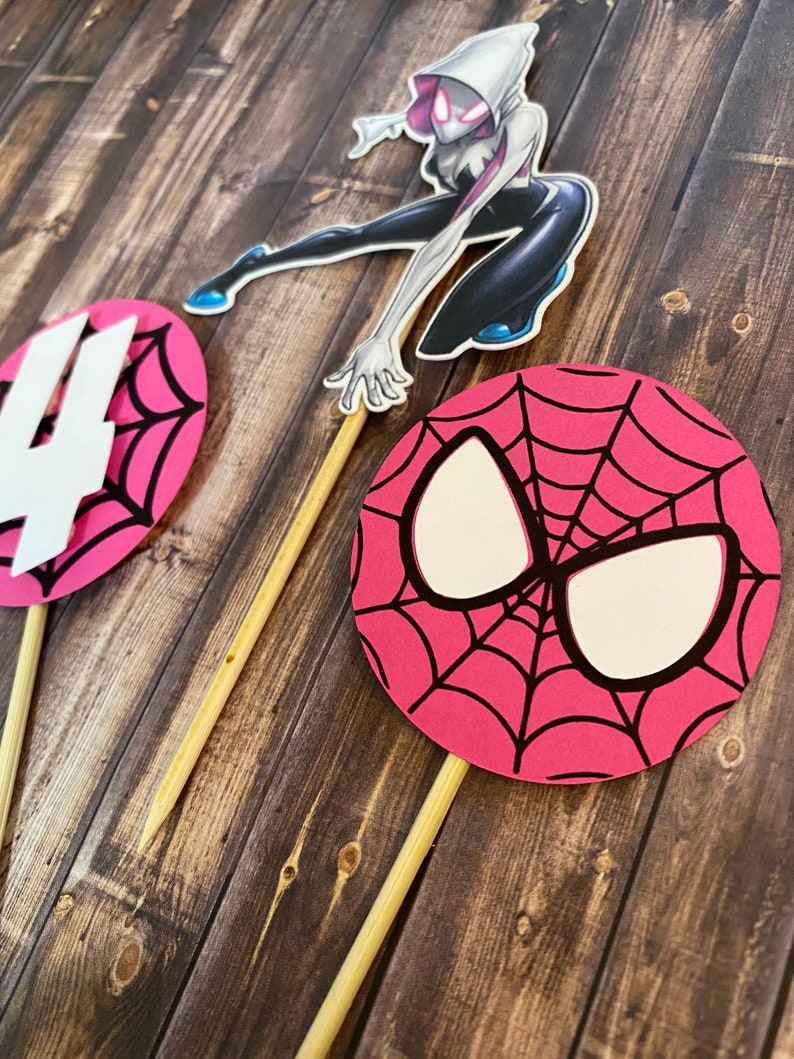 May include: Three pink and black Spider-Man themed cake toppers. One topper features a white Spider-Gwen character, another has a pink and black Spider-Man mask, and the last topper is a pink and black web design with a white number four.