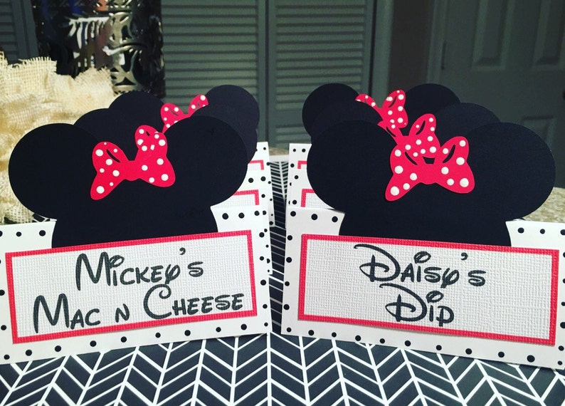 May include: Two white cardstock food labels with red borders and black polka dots. Each label features a black silhouette of Minnie Mouse's head with a red bow with white polka dots. The text on the labels reads "Mickey's Mac N Cheese" and "Daisy's Dip".