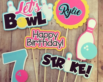 May include: A collection of bowling-themed party decorations on wooden sticks. The decorations include the phrases "Let's Bowl," "Happy Birthday!," and "Strike!" along with a number 7, a name, bowling pins, and bowling balls. The color scheme is pink, teal, yellow, and black.