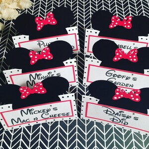 May include: Six black and red Mickey Mouse themed food labels with white polka dots. The labels have the names "Pluto's", "Minnie's", "Mickey's Mac N Cheese", "Goofy's Garden", "Daisy's Dip", and "Isabelle's".
