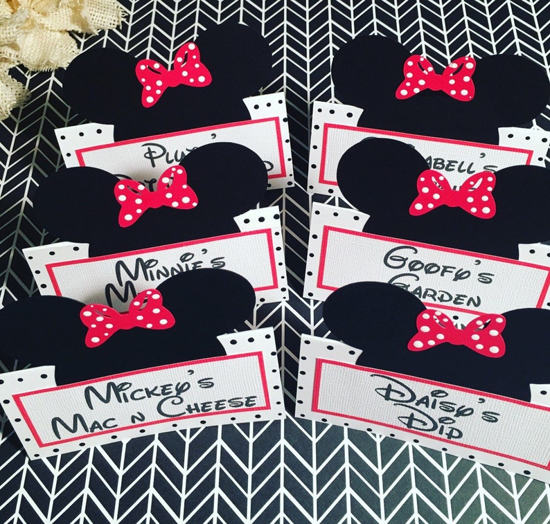 May include: Six black and red Mickey Mouse themed food labels with white polka dots. The labels have the names "Pluto's", "Minnie's", "Mickey's Mac N Cheese", "Goofy's Garden", "Daisy's Dip", and "Isabelle's".