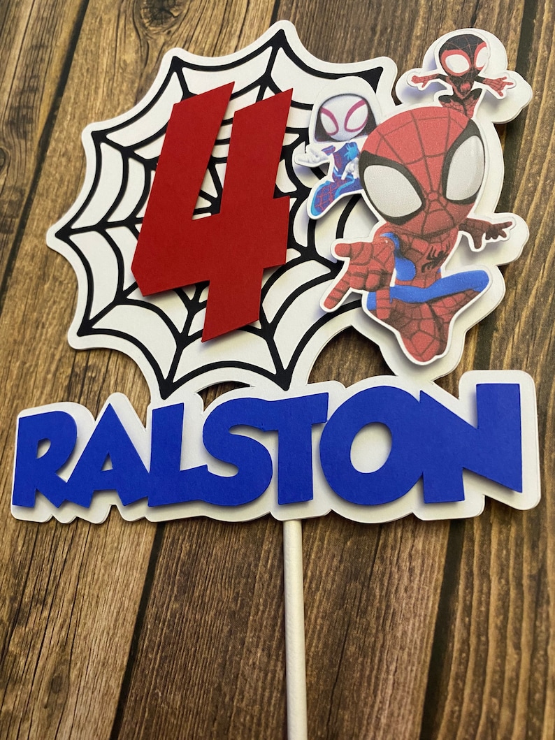 May include: A white cake topper with a red number 4, a black and white spiderweb, and three Spiderman characters. The name Ralston is written in blue letters.