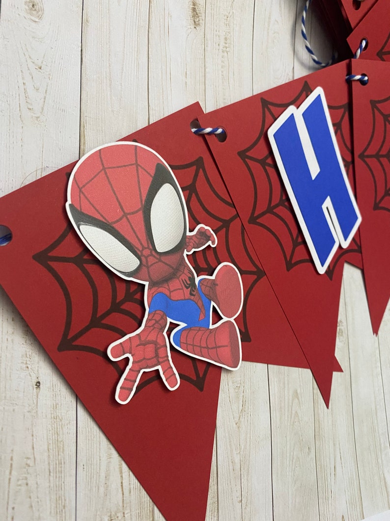 Puede incluir: Bandera de cumplea&ntilde;os de Spiderman roja y azul con un personaje de Spiderman y una letra H azul. La bandera est&aacute; hecha de cartulina roja con un dise&ntilde;o de telara&ntilde;a negra.