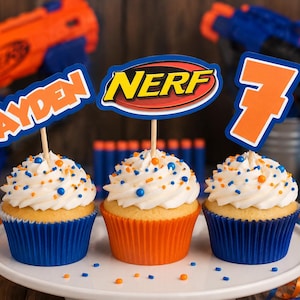 May include: Three cupcakes with white frosting and blue and orange sprinkles in blue and orange paper liners. Each cupcake has a wooden pick topper: one says AYDEN in orange, one has the NERF logo, and one is the orange number 7 with a blue outline.
