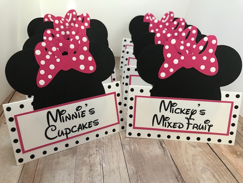 May include: Black and white polka dot table cards with a pink Minnie Mouse head cutout. The cards have the text "Minnie's Cupcakes" and "Mickey's Mixed Fruit".