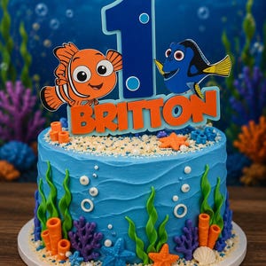 May include: A round blue cake decorated with edible sea life, including orange and blue starfish, coral, and white pearls. A cake topper features a large blue number one, an orange clownfish, a blue tang fish, and the name BRITTON in orange.