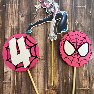 May include: Three cake toppers featuring Spider-Man characters. The first topper is a pink circle with a black spiderweb design and the number "4" in white. The second topper is a white and black illustration of Spider-Gwen. The third topper is a pink circle with a black spiderweb design and a white Spider-Man mask.