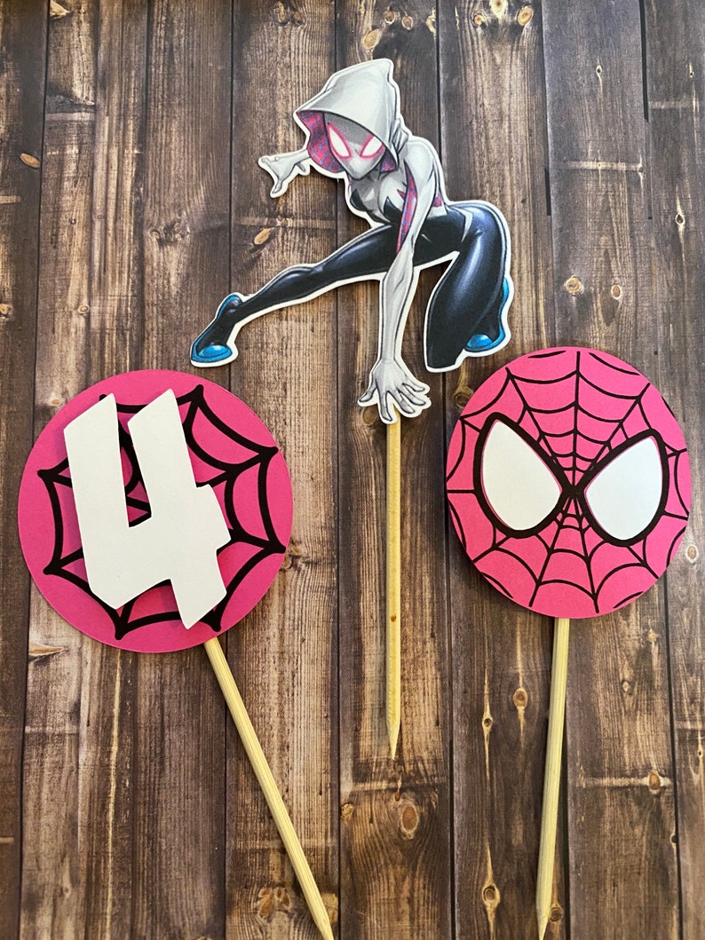 May include: Three cake toppers featuring Spider-Man characters. The first topper is a pink circle with a black spiderweb design and the number "4" in white. The second topper is a white and black illustration of Spider-Gwen. The third topper is a pink circle with a black spiderweb design and a white Spider-Man mask.