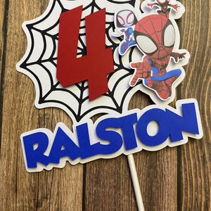 May include: A red number four, a black and white spider web, and three Spiderman characters on a white cake topper. The characters are Spiderman, Spider Gwen, and a black and red Spiderman. The cake topper also has the name Ralston in blue letters.