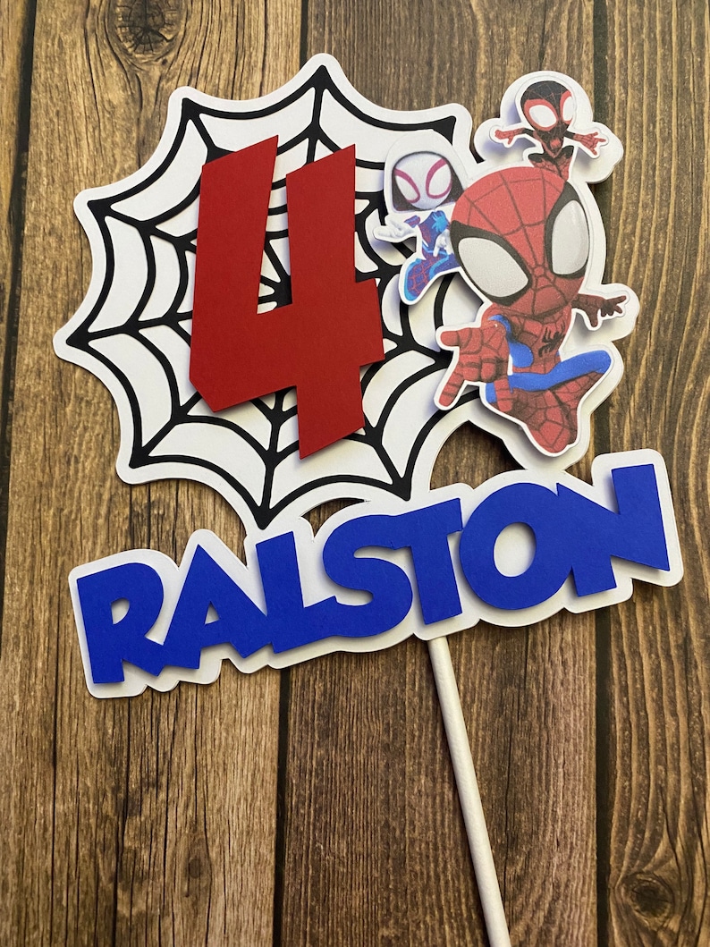 May include: A red number four, a black and white spider web, and three Spiderman characters on a white cake topper. The characters are Spiderman, Spider Gwen, and a black and red Spiderman. The cake topper also has the name Ralston in blue letters.