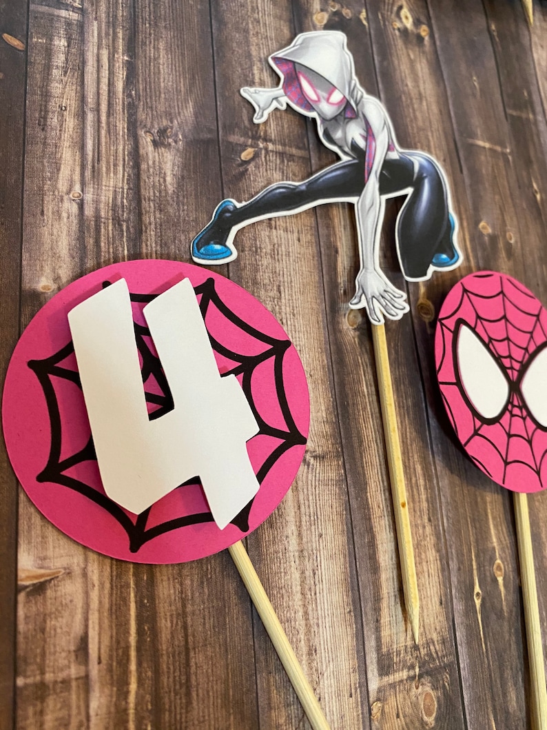 May include: Pink and black spiderweb cake topper with a white number 4 and a white spider-woman character.
