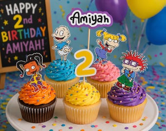 May include: A set of five cupcakes with orange, blue, yellow, pink, and purple frosting, topped with paper cutouts of cartoon characters, a yellow number two, and a purple sign that reads Amiyah, arranged on a white cake stand.