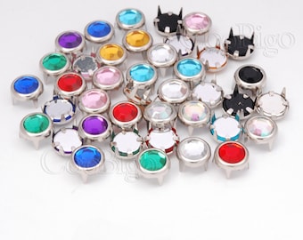 May include: Assortment of colorful round rhinestones with silver settings. The rhinestones are in various colors, including red, blue, green, yellow, pink, and clear. The rhinestones are set on a silver base with four prongs.