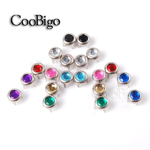 May include: Assortment of colourful round rhinestones with silver settings. The rhinestones are in various colours, including red, blue, green, purple, pink, and clear. The rhinestones are set on a white background.