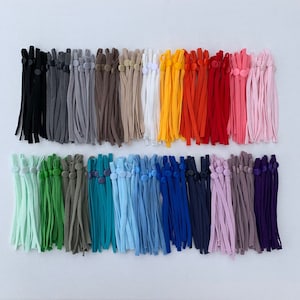 May include: A selection of adjustable face mask straps in a spectrum of colours, such as black, grey, brown, white, yellow, orange, red, pink, green, teal, blue, and purple. Each strap features a button for adjustment, neatly arranged on a white surface.