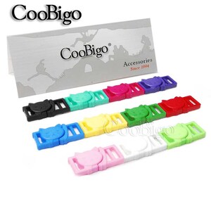 Op de afbeelding: Een assortiment van kleurrijke plastic kattenvormige schuifgespen. De gespen zijn in verschillende kleuren, waaronder zwart, groen, roze, geel, blauw, rood, wit en lichtgroen. De gespen zijn in een rij op een wit oppervlak gerangschikt. CooBigo Accessories Since 1994 is gedrukt op een witte kaart achter de gespen.
