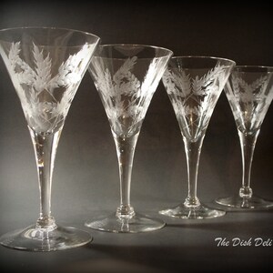 Op de afbeelding: Vier heldere glazen martini glazen met een gegraveerd ontwerp van tarwehalmen. De glazen zijn in een rij op een donkere achtergrond gerangschikt.