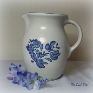 Może przedstawiać: Biały dzbanek ceramiczny z niebieskim wzorem kwiatowym. Dzbanek ma szeroki otwór i uchwyt. Wzór kwiatowy to stylizowany niebieski kwiat z zieloną łodygą.