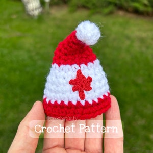 May include: A small crocheted hat in red and white with a red maple leaf design. The hat is a miniature version of a Canadian Santa hat.  The text "Crochet Pattern" is visible below the hat.