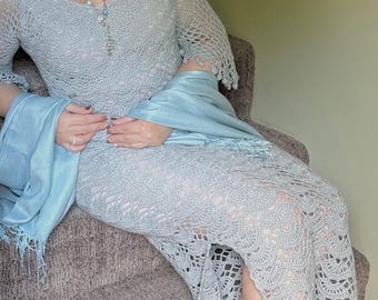 May include: A light blue crochet dress with long sleeves and a V-neck. The dress has a detailed openwork pattern and a slit on the side. A matching blue shawl is draped over the arms. A necklace with blue and white beads is also visible.