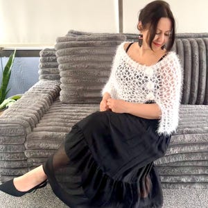 May include: A woman wearing a white, open-weave, fuzzy sweater and a long, black skirt. She is seated on a gray, textured sofa, wearing black flats. The outfit is a mix of textures and styles.