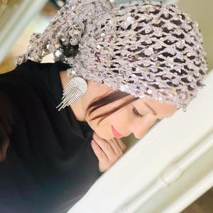 Puede incluir: Un cubrecabezas de crochet plateado con lentejuelas con un diseño suelto y fluido. El cubrecabezas está hecho de una tela delicada de tejido abierto con pequeñas lentejuelas plateadas en todo.