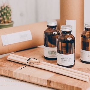 May include: Three brown glass bottles with silver lids and labels containing the text "Starry Lagoon", "Midnight Meadow", and "Midsummer Blossom". The bottles are sitting on a wooden chopping board with two wooden sticks and a black string with a tag that says "Do you love you?" The bottles are in front of two brown cardboard tubes.