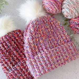 May include: Two handmade knit beanies with white faux fur pom-poms. One beanie is a deep red with multicolored flecks, and the other is a mix of purple, pink, and yellow. Yarn skeins in similar colors are also visible.