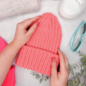 May include: A pink knitted beanie hat with a folded brim. The hat is being held by a person's hands.