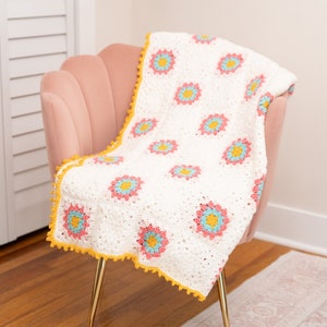 May include: A white crocheted throw blanket with a yellow border and a floral pattern in shades of pink, blue, and yellow. The blanket is draped over a pink velvet armchair with gold legs.