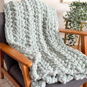 May include: A chunky knit blanket in a soft, muted green color is draped over a gray armchair with wooden arms. The blanket's texture is prominent, with large, looped stitches. The chair is set in a well-lit room, suggesting a cozy, inviting atmosphere.