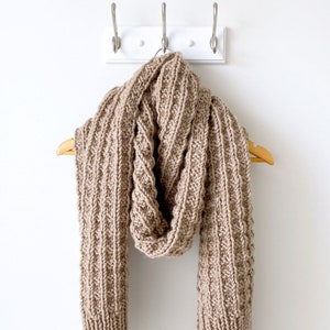 May include: A brown knitted scarf with a cable knit pattern hangs on a white hook.