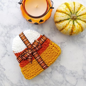 Può includere: Un cappello a maglia a forma di mais dolce con strisce arancioni, gialle e bianche. Il cappello è decorato con un fiocco a quadri. Nell'immagine sono presenti anche un portacandele a forma di zucca e una piccola zucca gialla.
