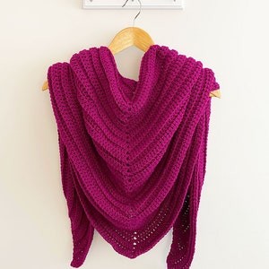May include: A magenta crocheted shawl with a v-shaped neckline, hanging on a wooden hanger.