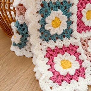 May include: Close-up of a handmade crochet blanket featuring daisy motifs. The blanket is composed of square blocks in white, pink, teal, and yellow. The daisy centers are yellow, with white petals and teal leaves. The blanket is on a wooden surface.