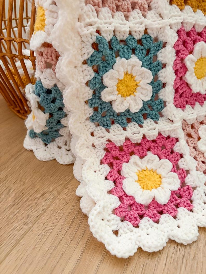 May include: Close-up of a handmade crochet blanket featuring daisy motifs. The blanket is composed of square blocks in white, pink, teal, and yellow. The daisy centers are yellow, with white petals and teal leaves. The blanket is on a wooden surface.