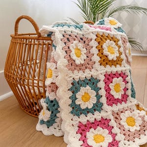 May include: A handmade crochet blanket with daisy flower squares in shades of pink, teal, yellow, and white. The blanket is folded over a wicker basket, creating a cozy and inviting scene. The blanket's edges have a scalloped trim.
