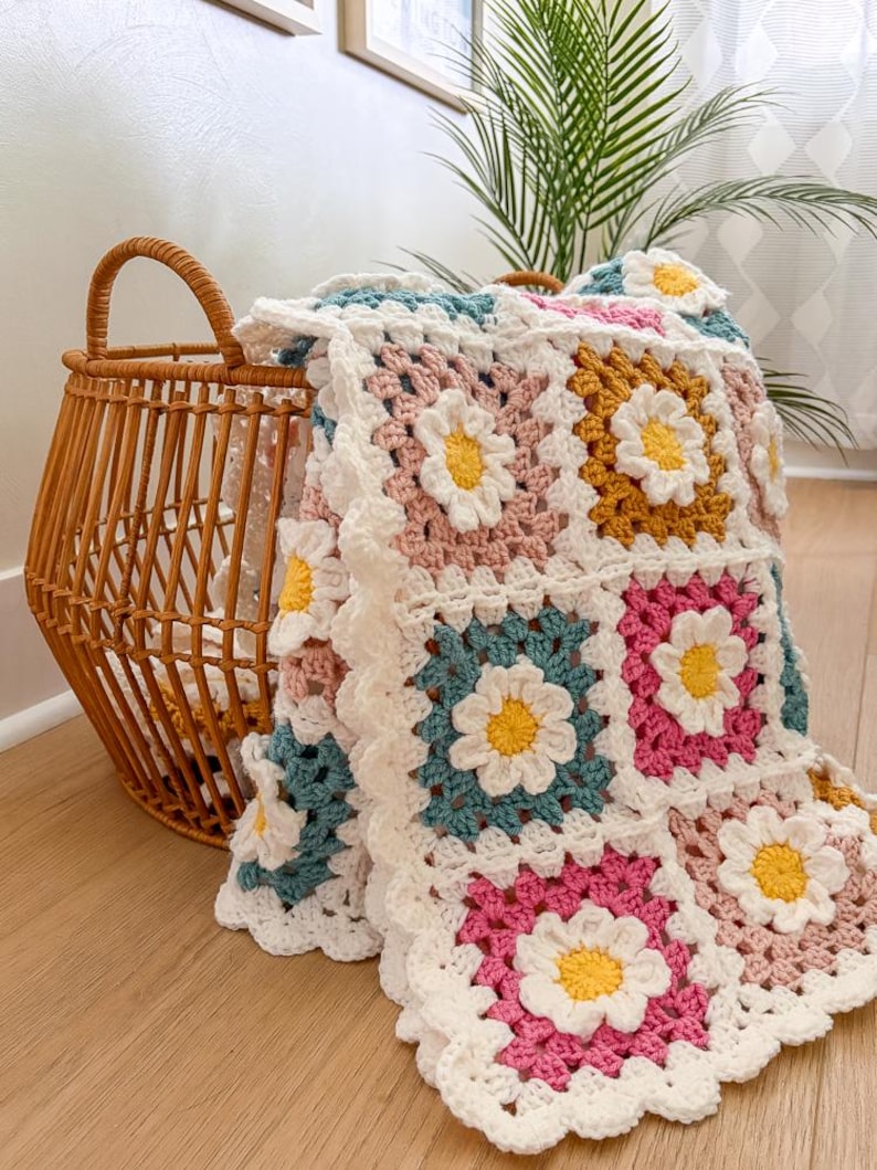 May include: A handmade crochet blanket with daisy flower squares in shades of pink, teal, yellow, and white. The blanket is folded over a wicker basket, creating a cozy and inviting scene. The blanket's edges have a scalloped trim.