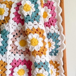May include: A handmade crochet blanket featuring a daisy flower design. The blanket incorporates white, yellow, pink, teal, and gold squares. The edges are scalloped, adding a decorative touch to the cozy, colorful blanket.